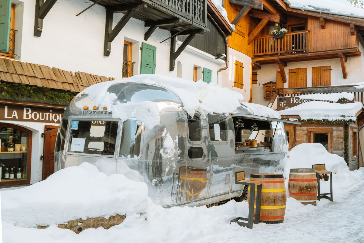 An Airstream trailer, covered in snow, parked outside a restaurant. There are barrels set up as tables in a small dining area beside the trailer