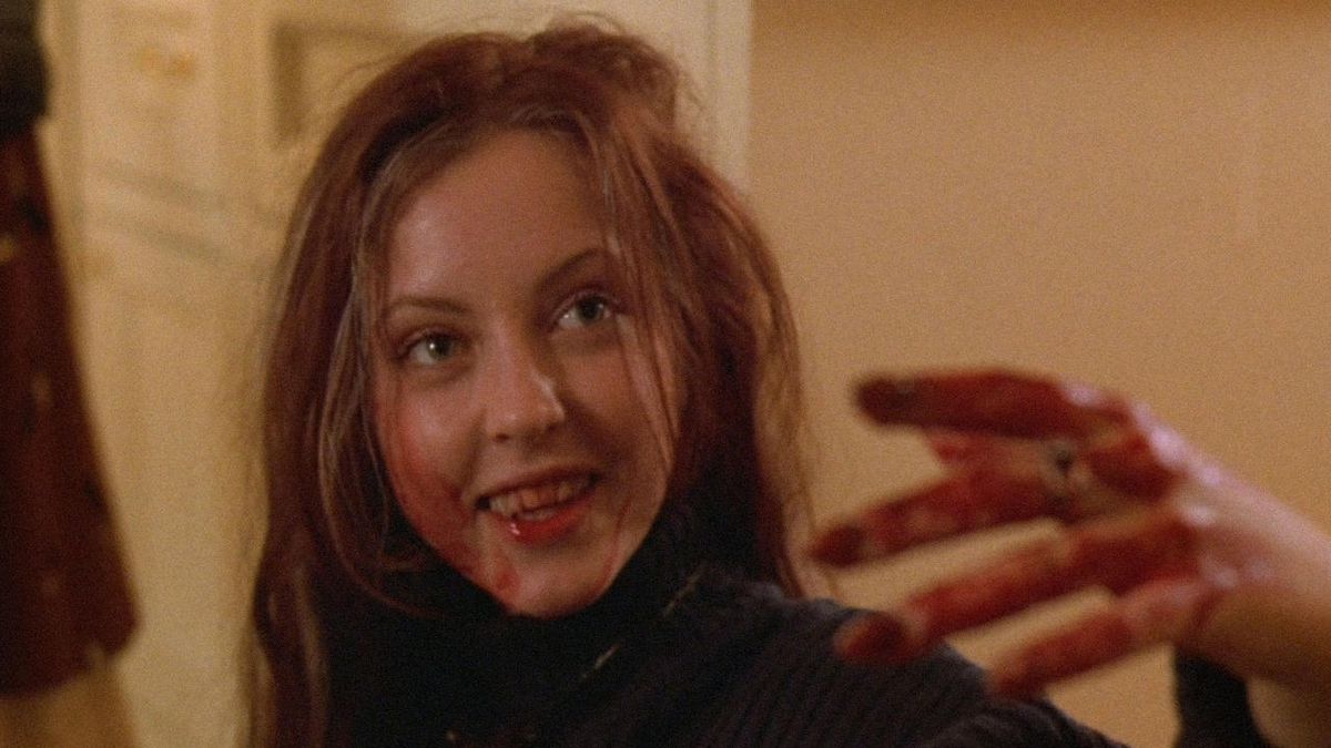 A woman with blood on her face holds her bloody hand up, out of focus, with a smile on her face in Ginger Snaps