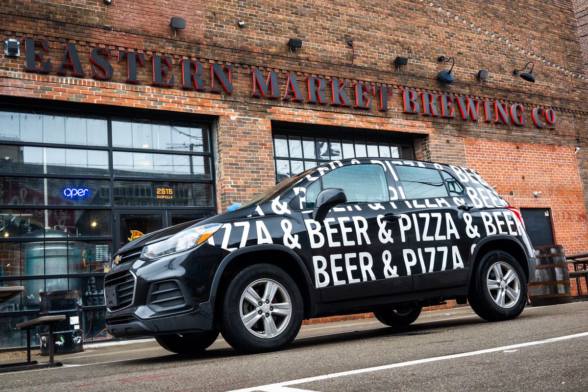 A black vehicle with white print on it that says pizza &amp; beer repeatedly parked on a street in front of a brick building that says Eastern Market Brewing Co.