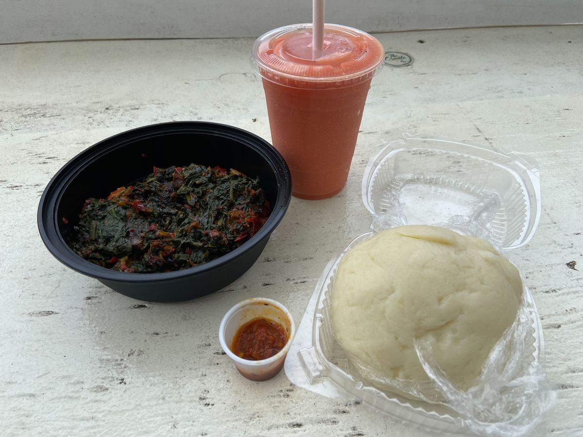 A black takeout container holds a spinach-based stew, pictured next to a pink smoothie, and a clear plastic container holding a ball of pounded white yam.