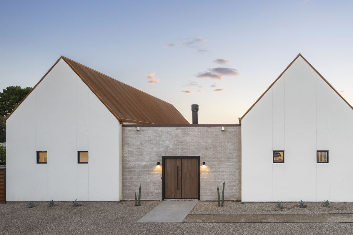 Exterior shot of home formed by two gabled structures connected by a low brick wall in a desert landscape. 