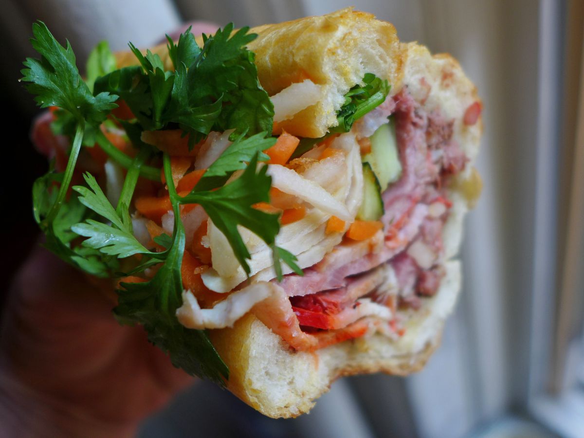 A bright banh mi sandwich seen in cross section with orange carrots, leafy deep green sprigs of cilantro, and layered meats.