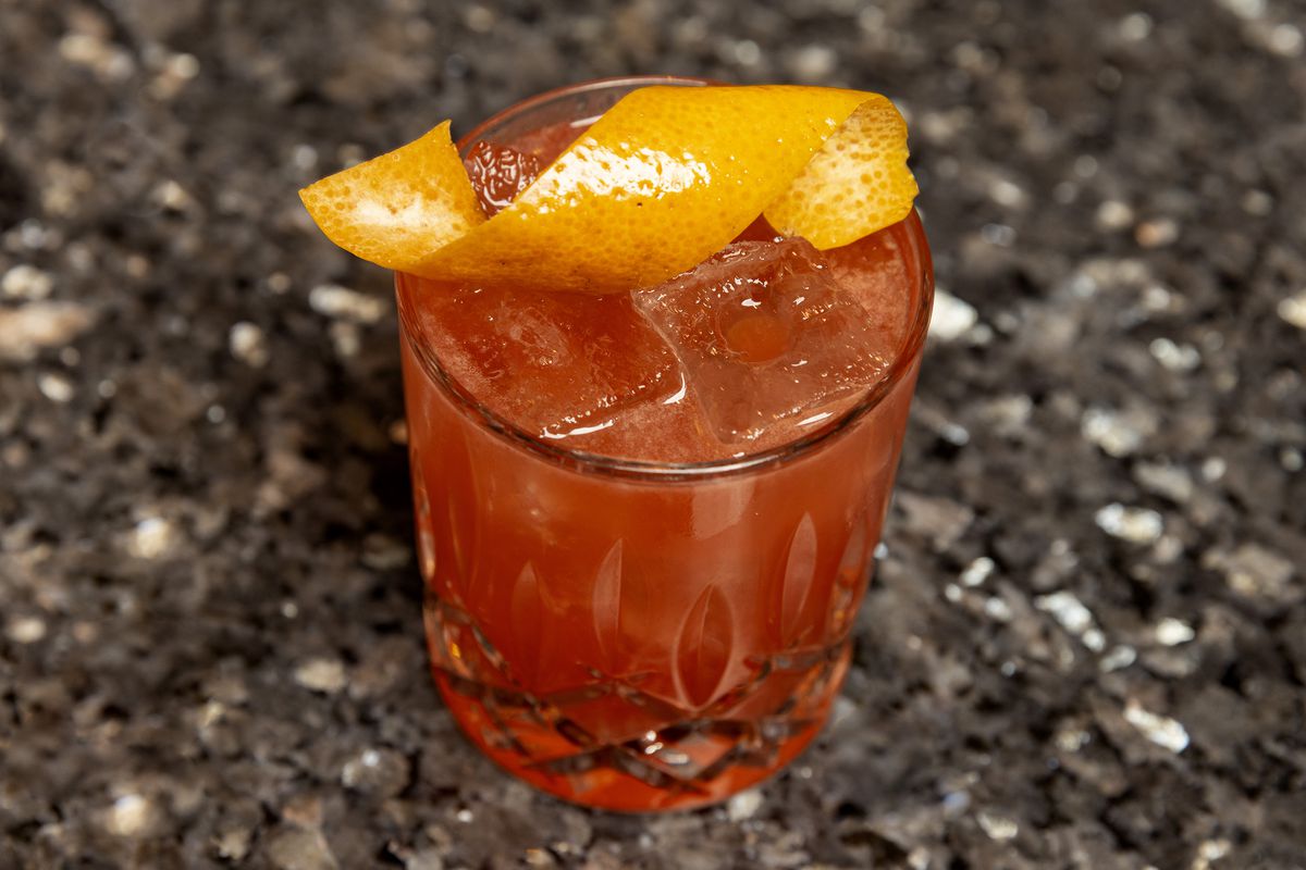 A red-pink cocktail in a rocks glass with an orange peel twist garnish.