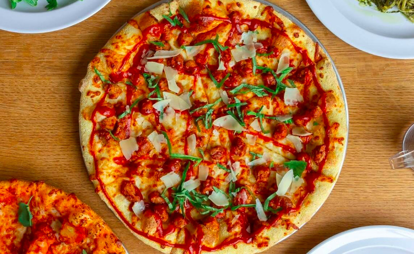 A gluten-free vegetable pizza drizzled with hot sauce sits atop a tray on a wooden table. 