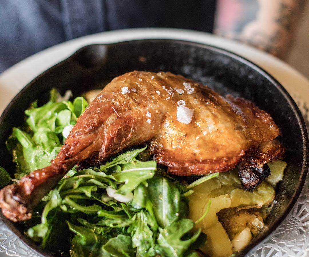 A roasted chicken leg in a cast iron pan on a bed of greens and vegetables.