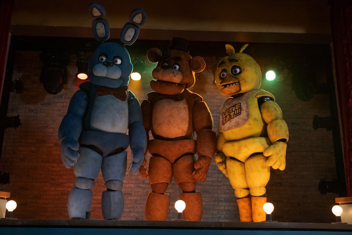 Animatronics Bonnie, Freddy Fazbear, and Chica stand on a stage, looking to the right, in the Universal Pictures movie adaptation Five Nights at Freddy’s
