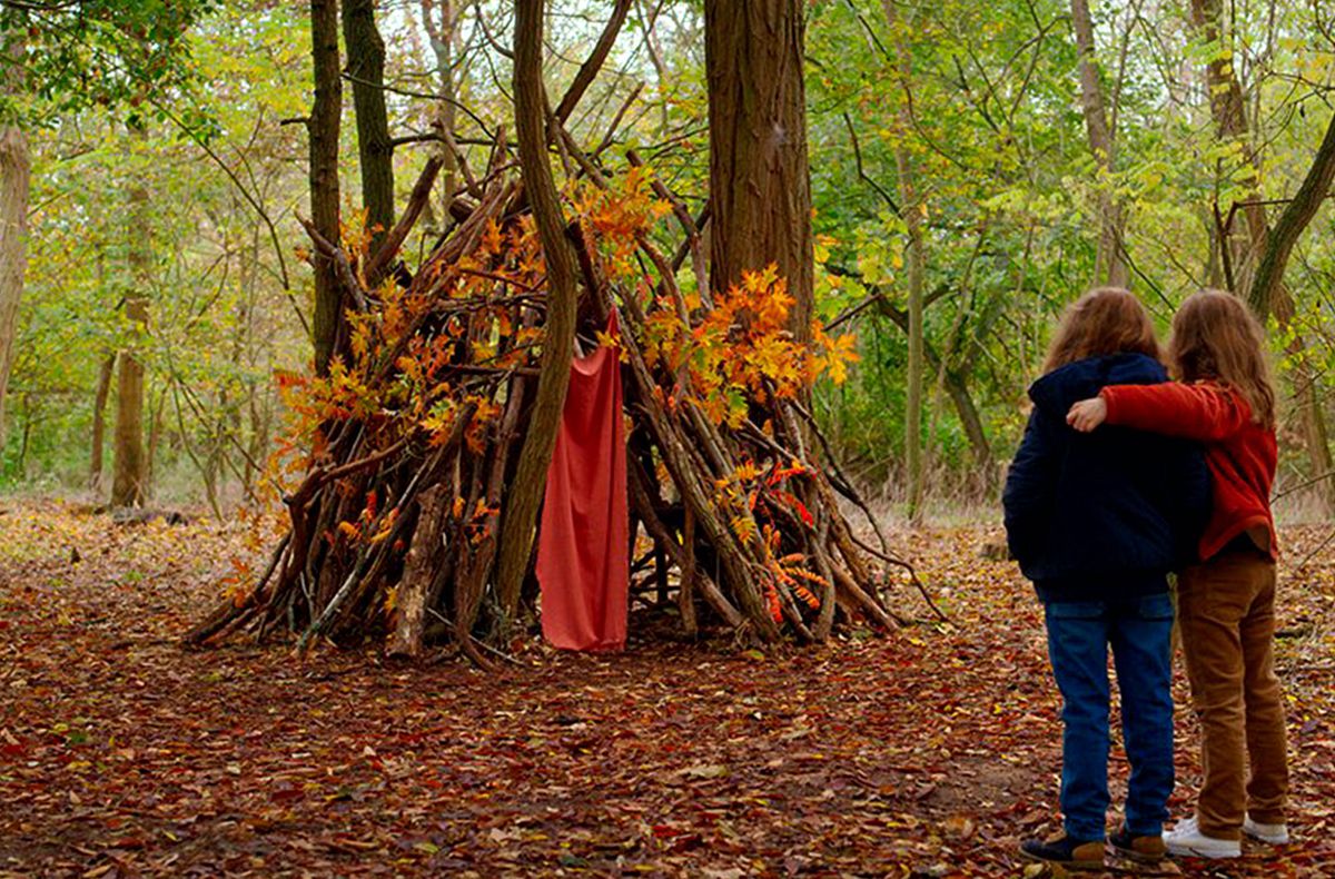 Two young girls with their arms around each other look at their stick fortress in the forest in Petite Maman