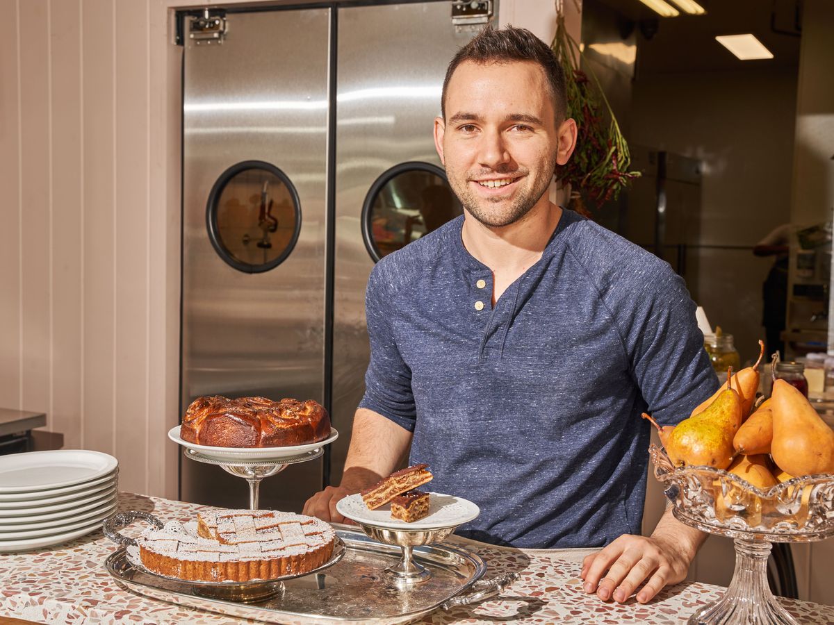 Executive chef and owner of Agi’s Counter, Jeremy Salamon stands behind a terrazzo counter topped with pastries on tiered trays, while smiling and wearing a blue shirt. 