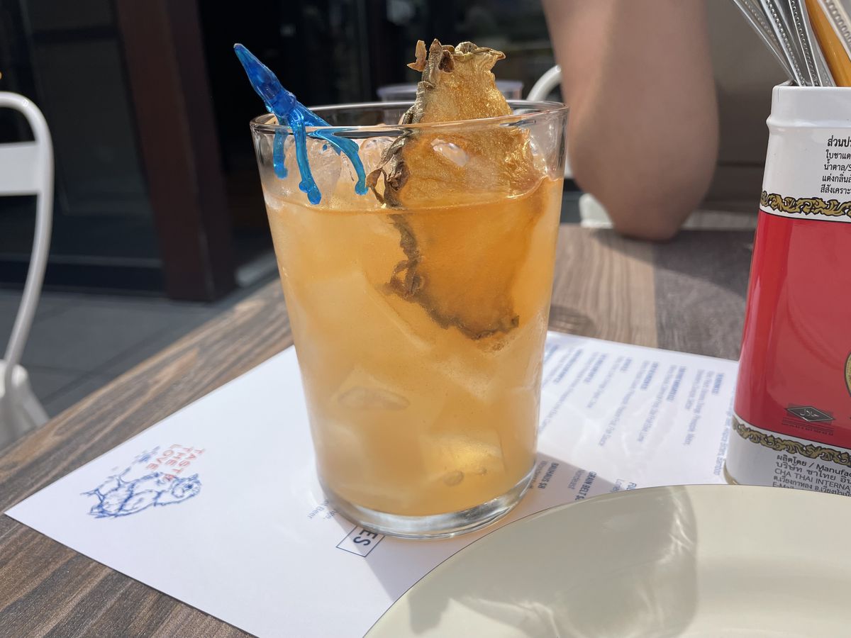 A light orange colored cocktail with a slice of dried pineapple in it on a white menu on a wooden table. 