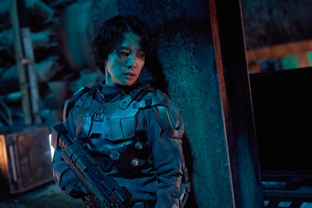 Eine Frau in futuristischer Rüstung versteckt sich hinter einer Betonsäule, die ein Gewehr hält.
