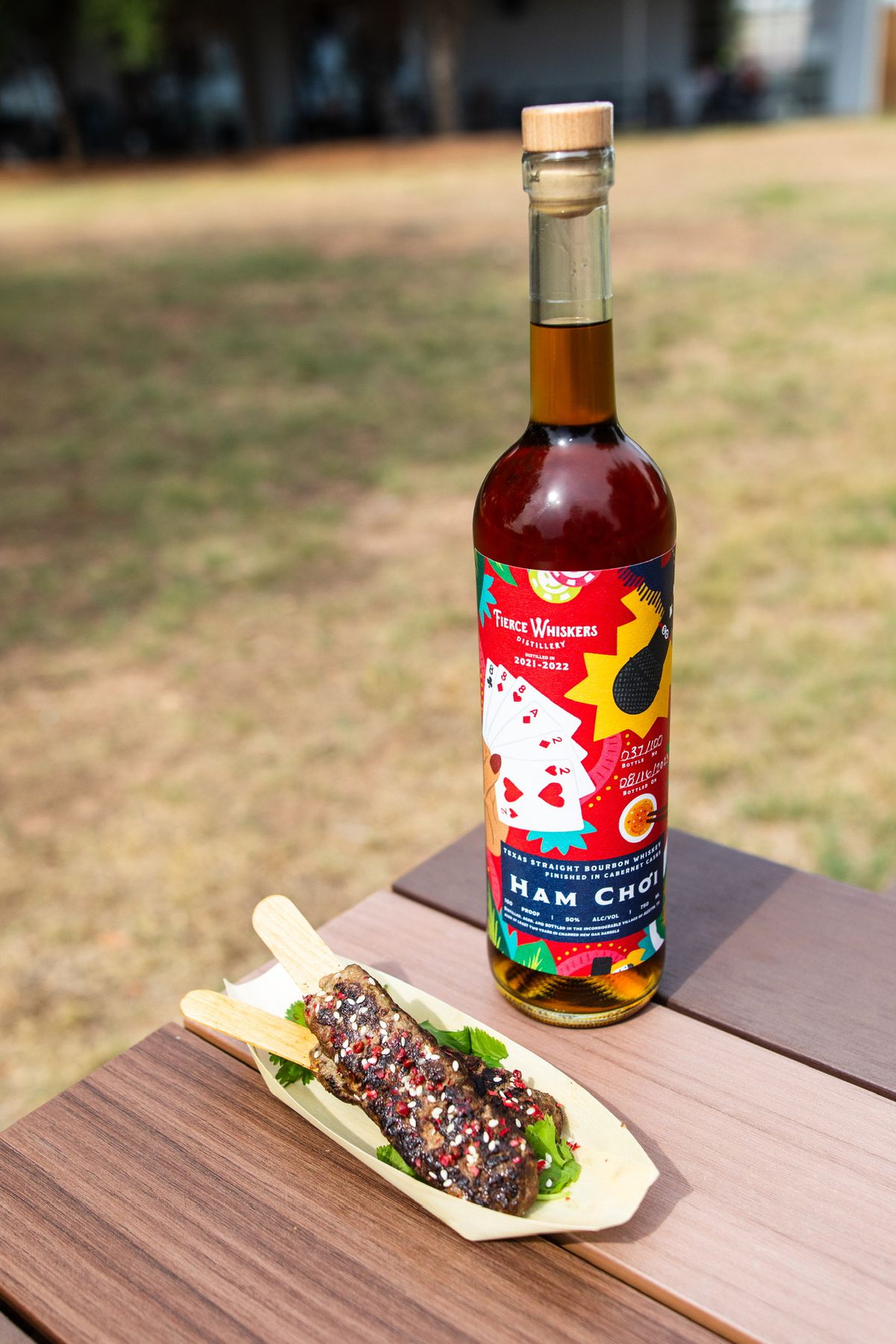 A bottle of whiskey with a bright label next to a meat kebab in a smaller tray on a picnic table outside. 