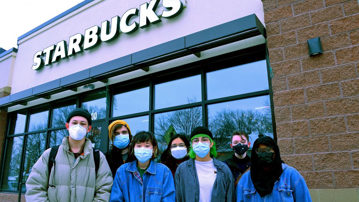 Seven Starbucks workers wearing jackets and face masks stand outside a Starbucks store with the logo prominently visible.