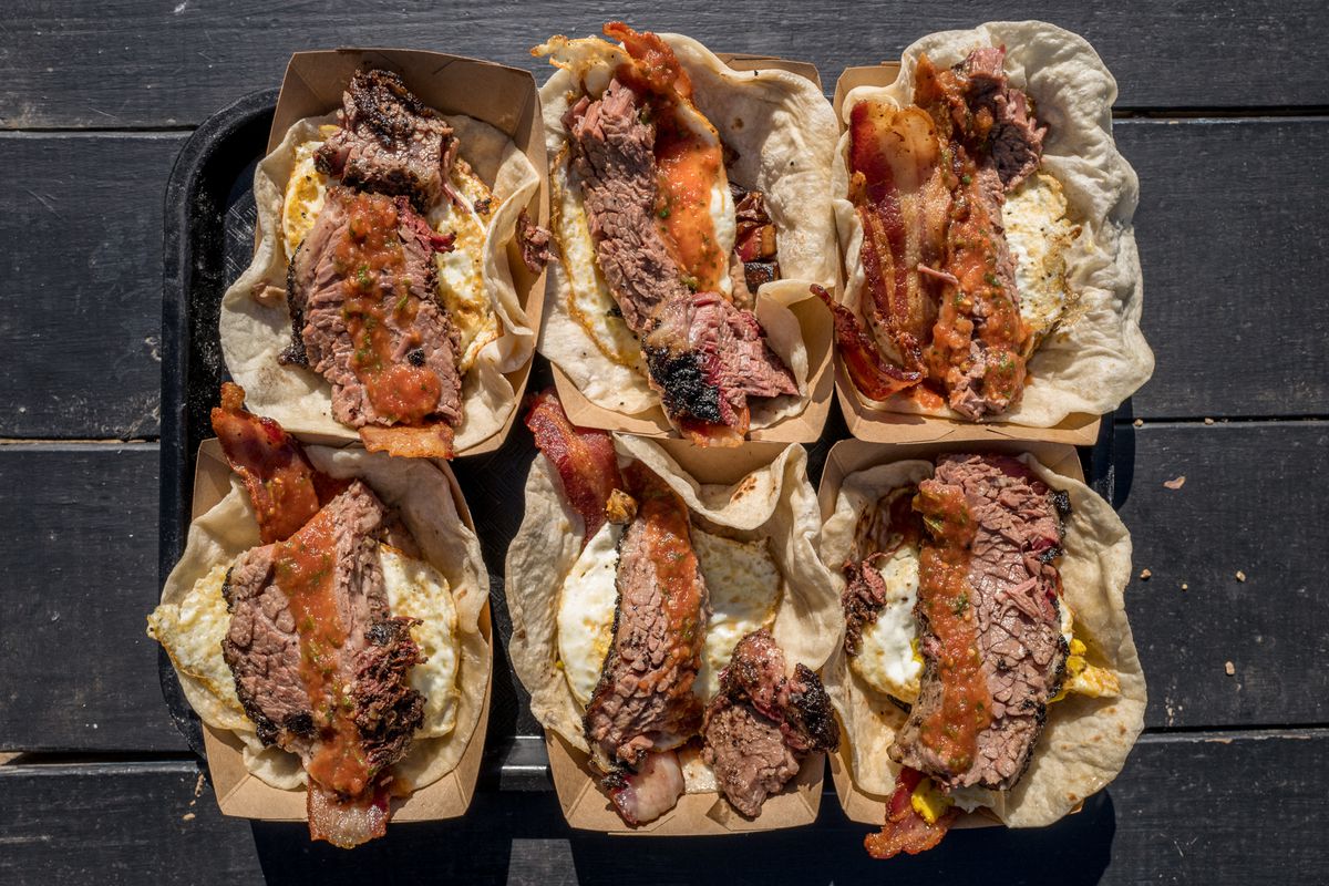 A tray of six tacos, each filled with a slice of bacon, brisket, and fried eggs.