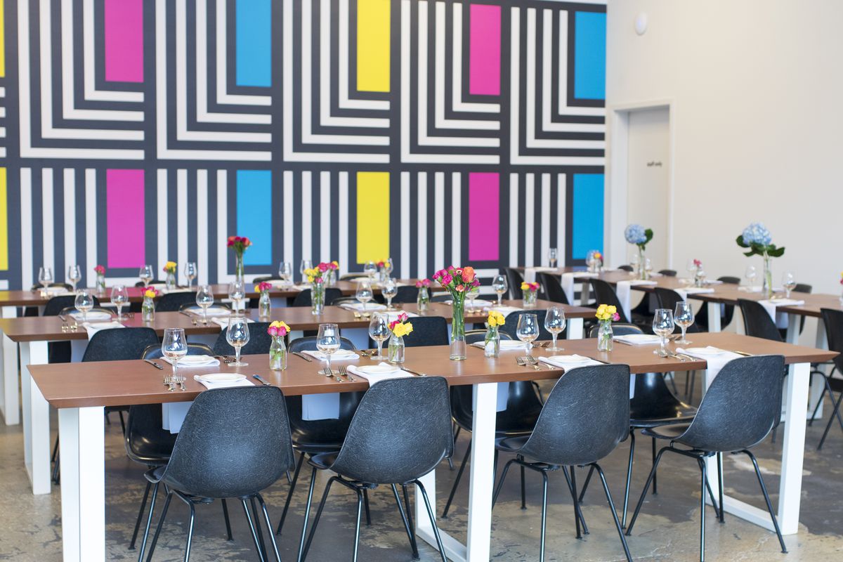 interior of aslin brewery with a colorful, abstract, geometric back-drop on the wall and black chairs situated around white tables.