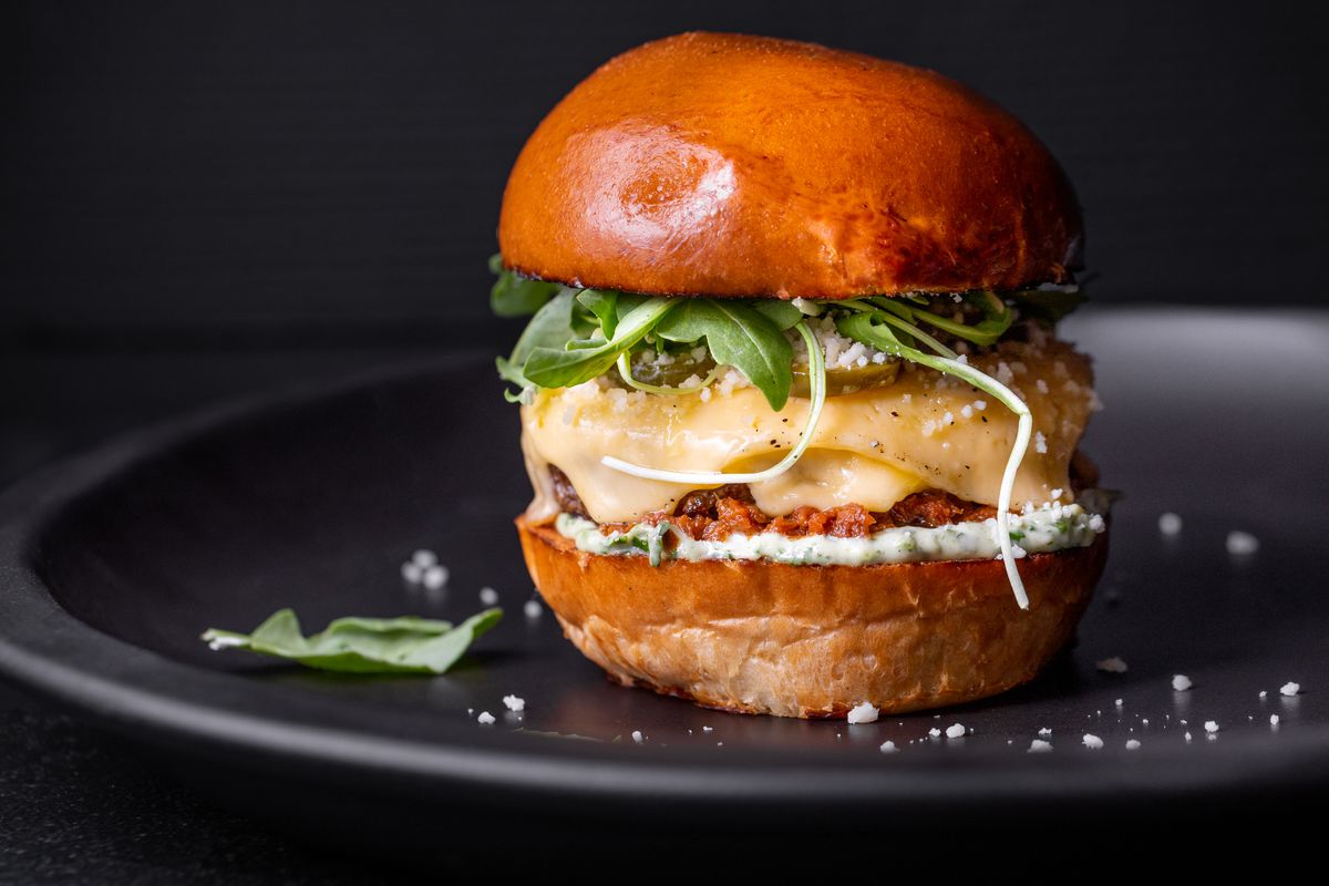 A tall burger on a golden brioche bun with lots of cheese and arugula.
