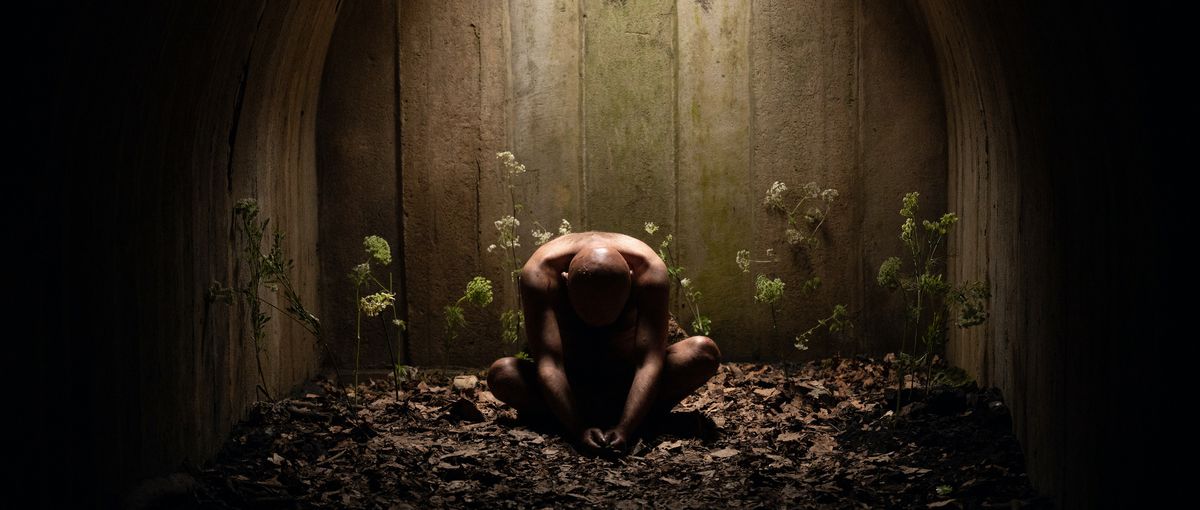 Rory Kinnear sits naked, hunched over, in a pile of dry leaves surrounded by stringy plants in Men