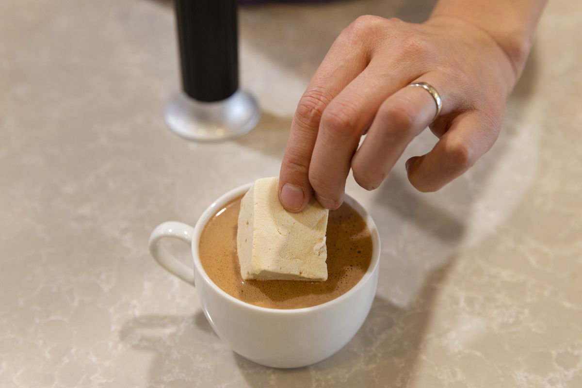 A hand places a marshmallow into a white mug of drinking chocolate.