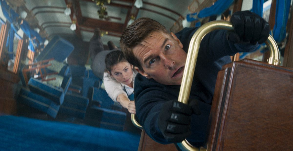 Tom Cruise as Ethan Hunt holds on to a railing in a train car turned vertical as Hayley Atwell clings on to him In Mission: Impossible — Dead Reckoning Part 1