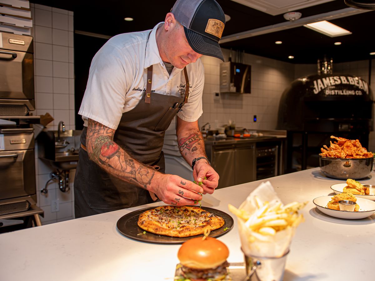 A chef prepares a thick flatbread dish on a kitchen counter beside other dishes in stages of prep. In the back is a large pizza oven branded James B. Beam Distilling Co