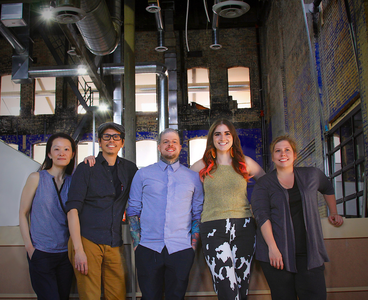 Five people stand in a line in a high-ceilinged industrial building. Their arms are around each other and they are smiling. 
