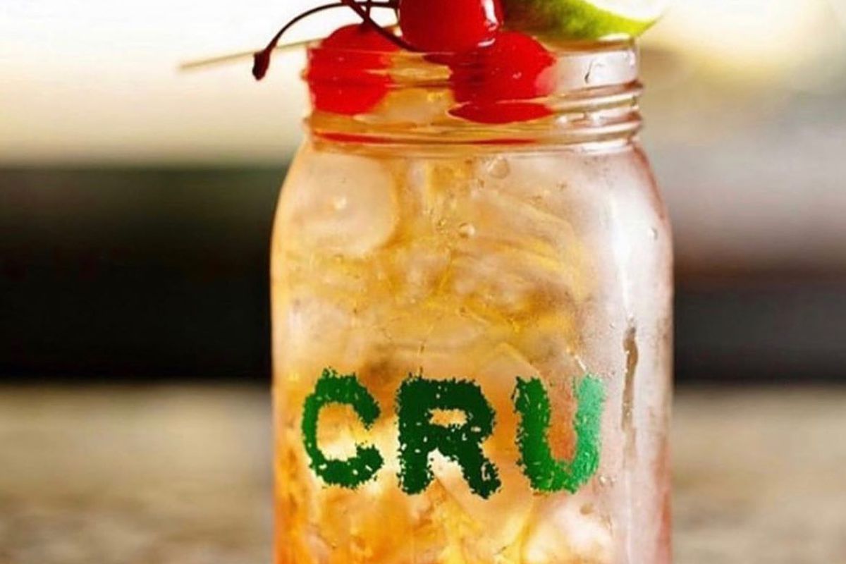 a glass jar with green letters that say “cru” on it, filled with an amber-colored cocktail and garnished with cherries and a lime