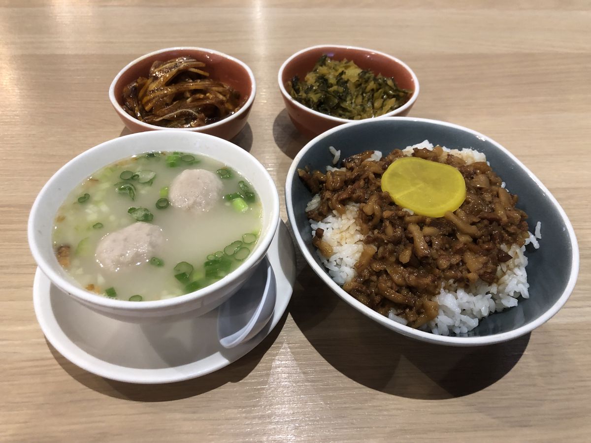 Four ceramic bowls on a light wooden surface holding diced braised pork belly over rice, a pork meatball soup with a milky broth, glistening chili-oil sliced pig ears, and pickled mustard greens. 