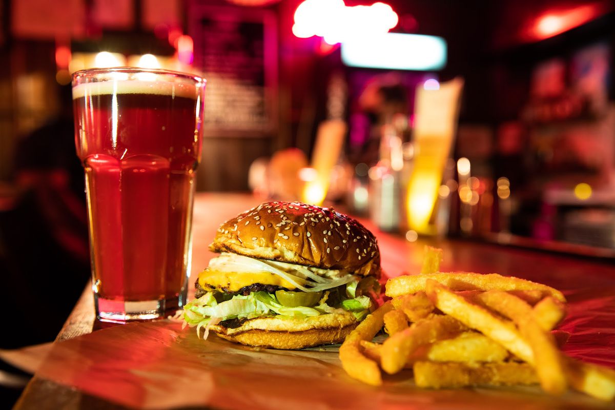 A glass of beer, a double cheeseburger, and a pile of fries on top of a bar with a blurred background