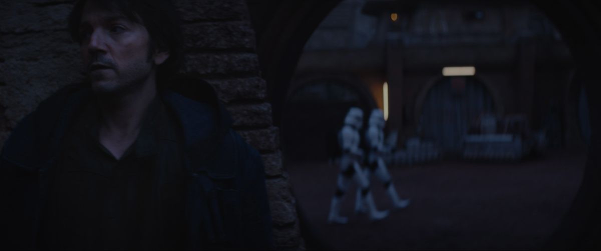 Cassian Andor hides behind a pillar at night while Stormtroopers march through the streets behind him