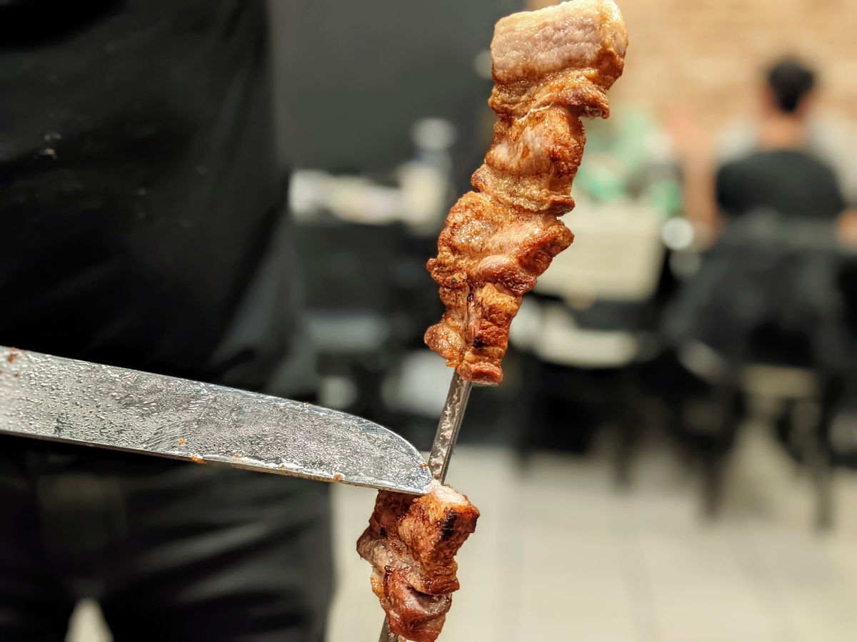 A knife cuts away at pork belly on a skewer inside of a restaurant.