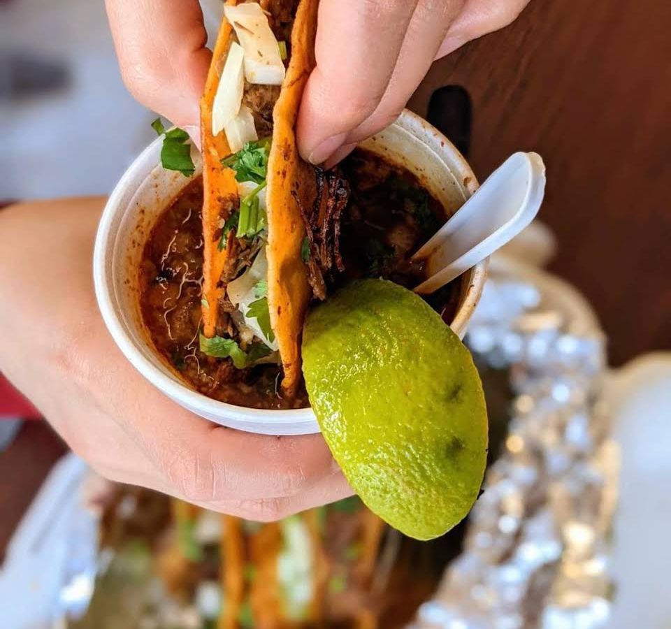 A person’s hands dip a birria taco into a Styrofoam cup of birria stem with a lime wedge on the rim and a plastic spoon.