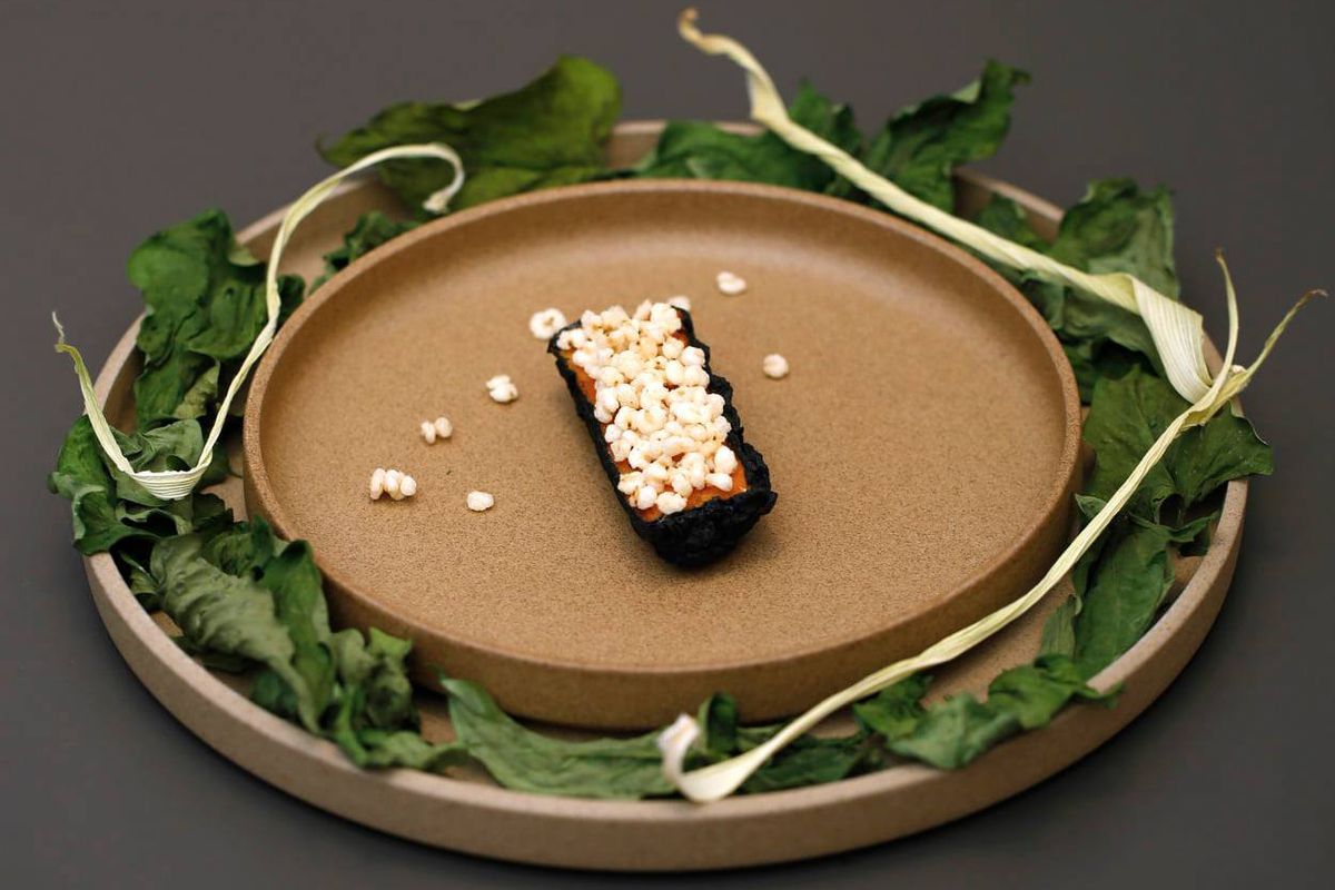 A piece of food, rectangular and topped with something resembling popcorn, at the center of a beige clay plate surrounded by leaves