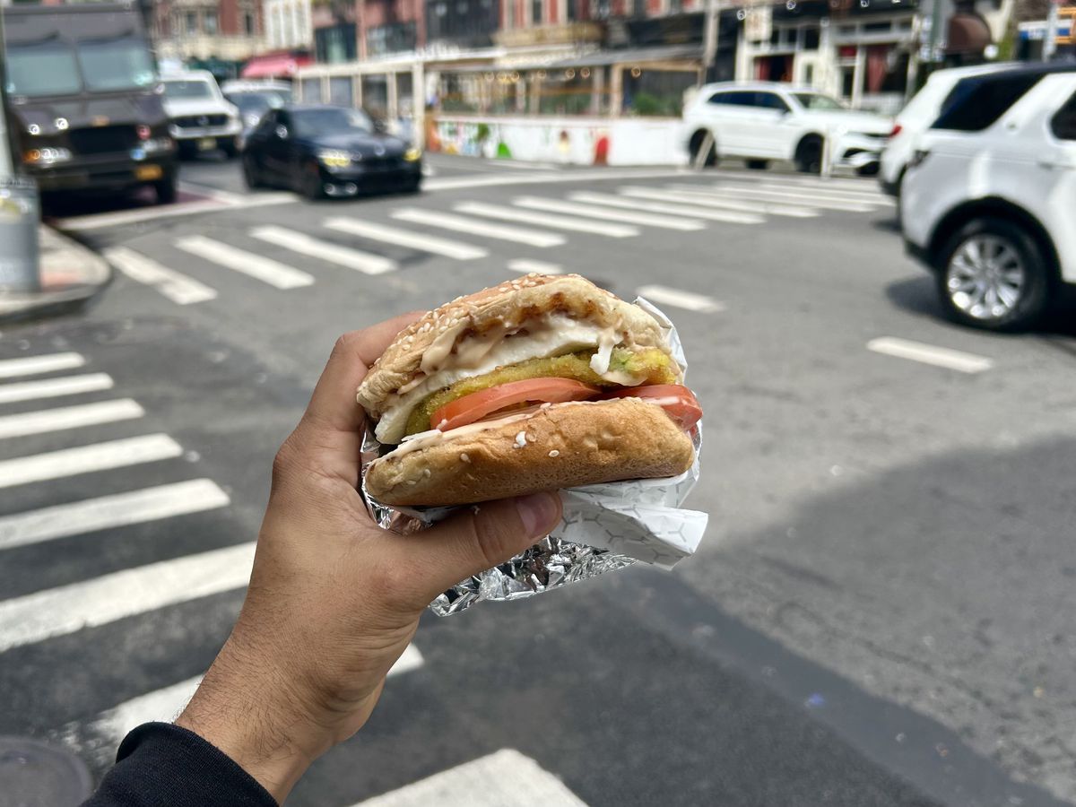 A hand with a black sleeve holds a burger with tomato, lettuce, and mayonnaise against a backdrop of a New York City street.
