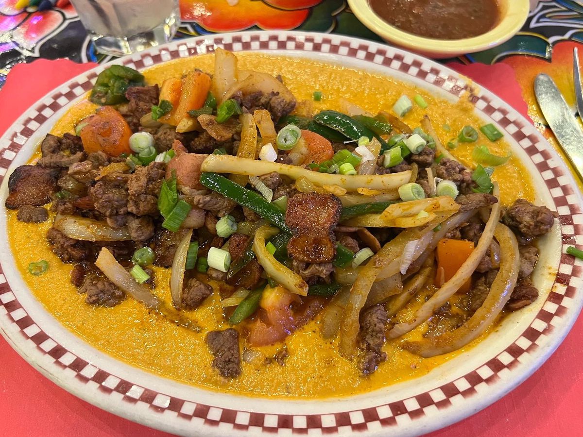 Slices of steak along with chopped bacon, jalapeno, onions, and tomatoes on a thick mole, served on a decorative plate with checkerboard pattern around the rim