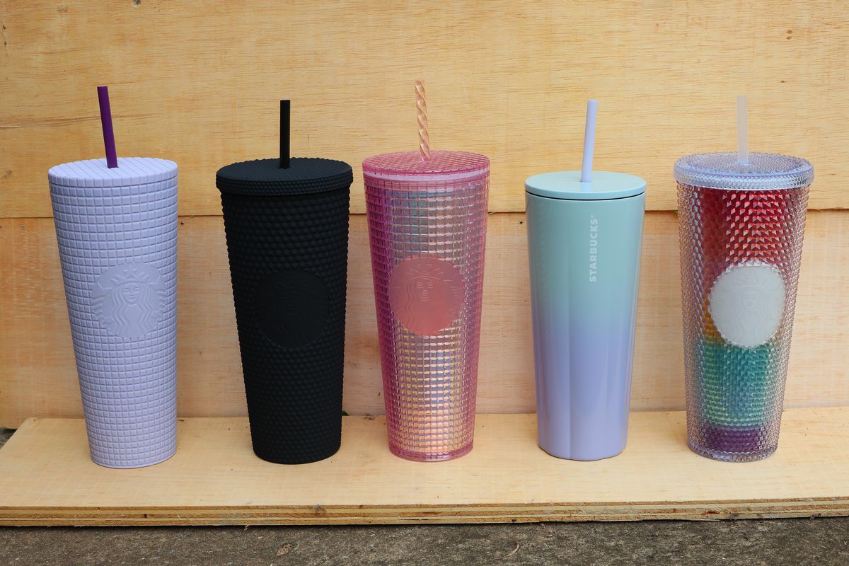 A row of reusable Starbucks cups on a plain wood shelf and background; from left to right, a tall lavender studded cup with a black straw, a tall black studded cup with a black straw, a tall pink iridescent studded cup with a pink straw, a tall ombre cup fading from light blue to lavender, and a tall rainbow studded cup with a clear top and straw.