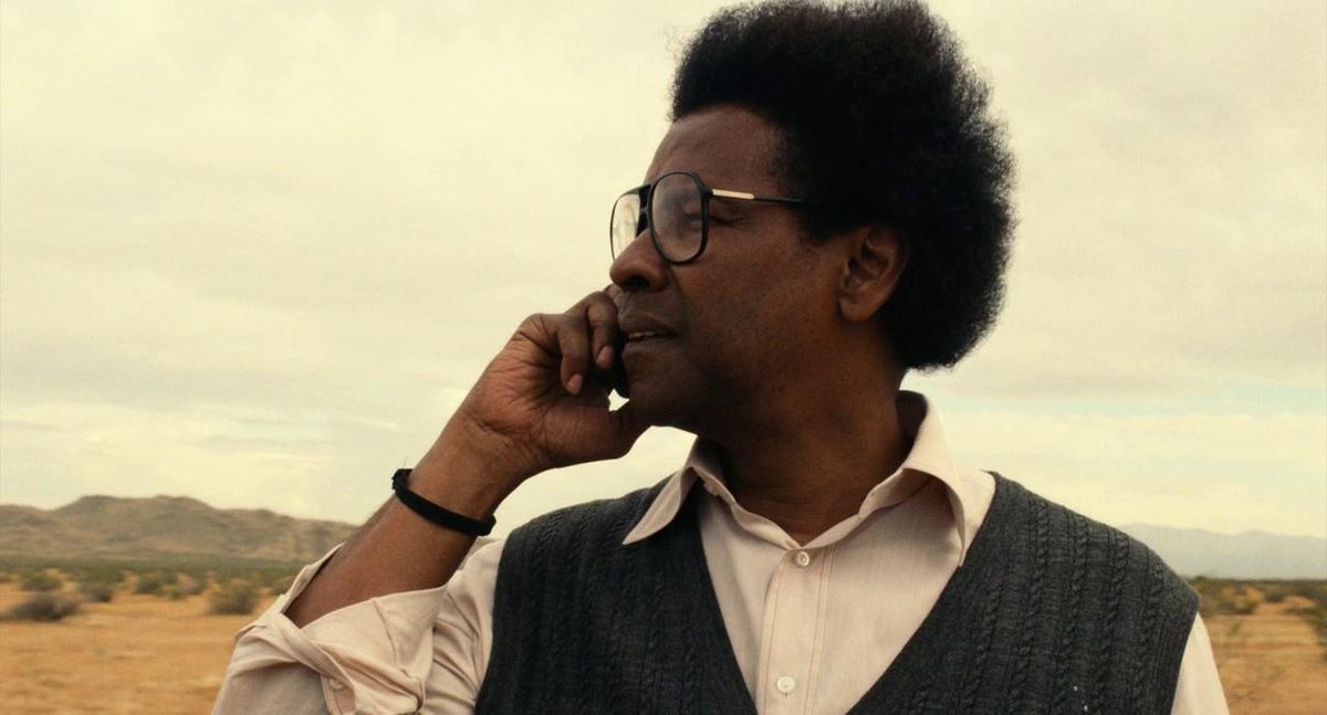 Denzel Washington holds his phone to his right ear while standing in the desert in Roman J. Israel, Esq. His hair is disheveled and he wears big glasses and a vest.