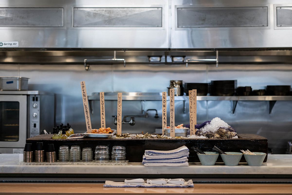 Tall wooden sticks showing names of raw seafood items, backed by a kitchen, at new restaurant Joyce.