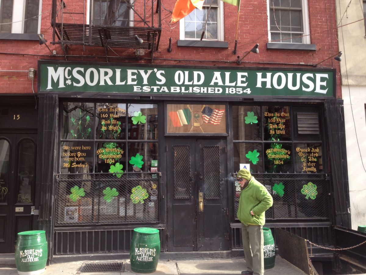 A man dressed in green walks in front of a restaurant decorated for Saint Patrick’s Day, with a sign that reads “McSorley’s Old Ale House”