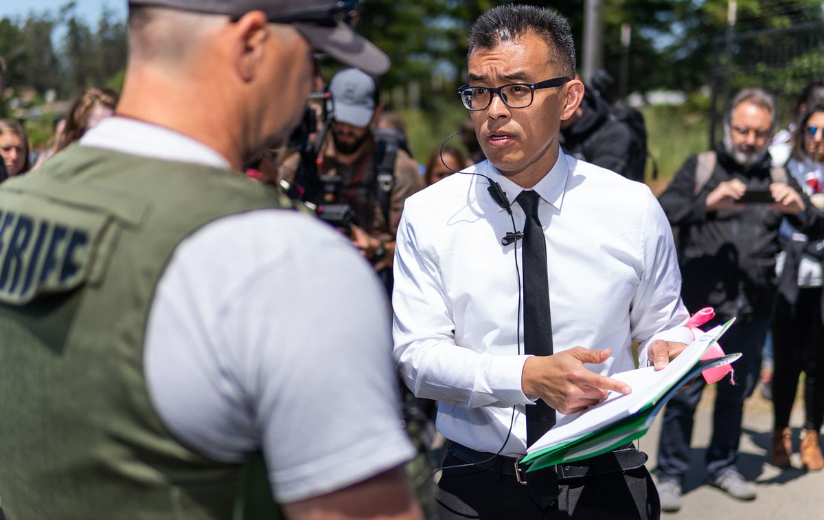 Hsiung in a business shirt and tie points to a stack of documents and talks to law enforcement on the street outside a farm.