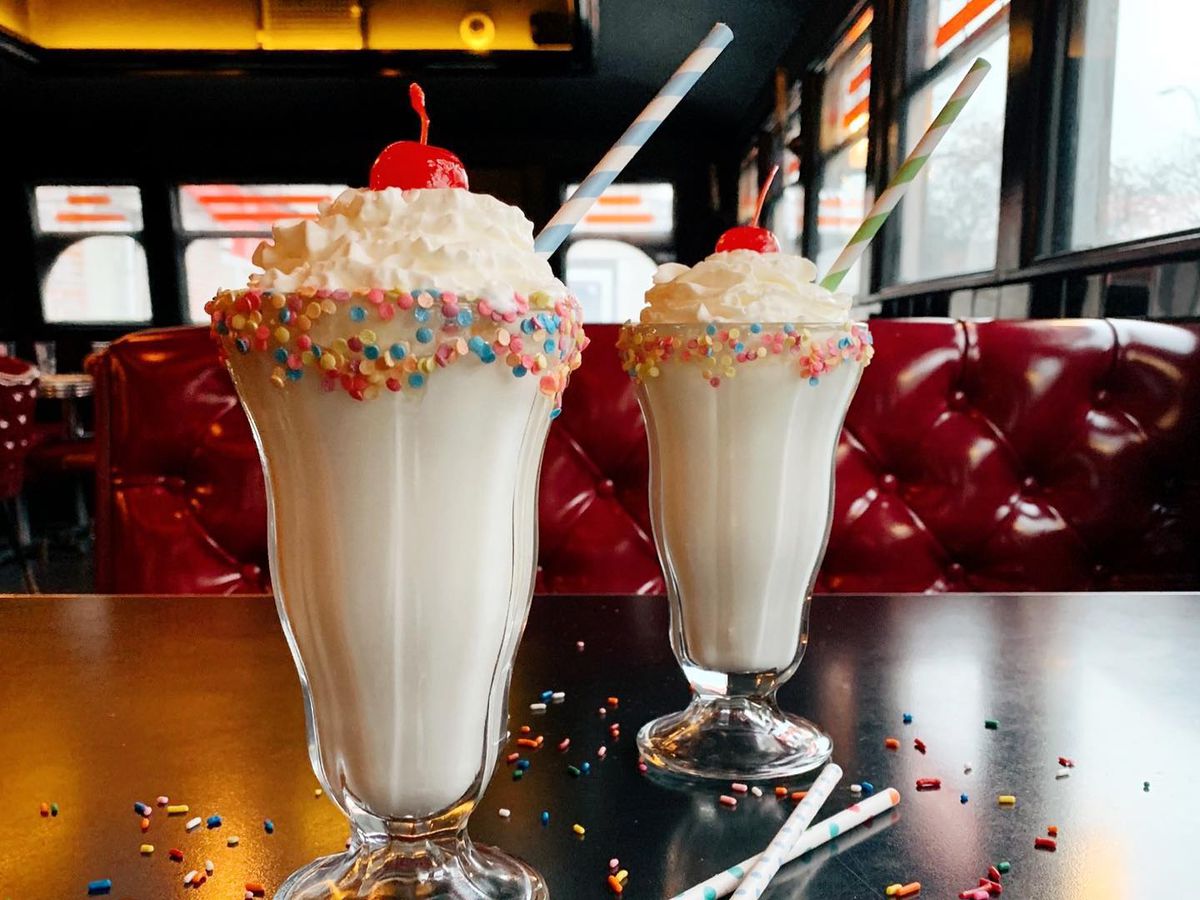 Two vanilla milkshakes with whipped cream, cherry, and rainbow sprinkles on the rim of the glass sit on a table in a shiny red booth in a sleek diner setting.