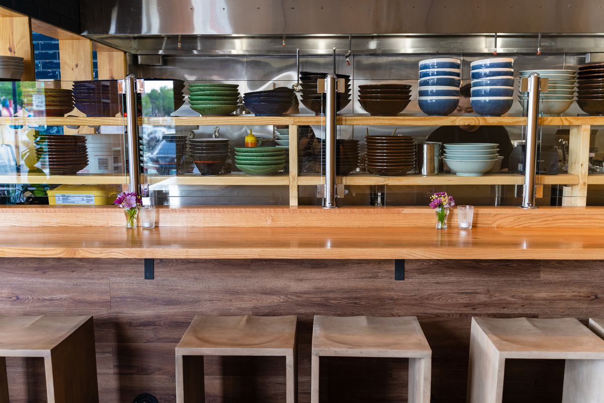A wooden ramen bar and kitchen with shelves piled with ramen bowls.