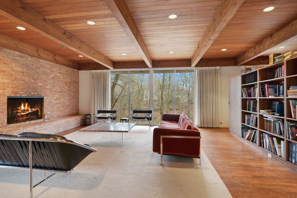 Interior shot of living room with beamed ceilings, brick hearth, floor-to-ceiling windows overlooking nature, and a sparsely decorated sitting area with modern furniture. A wooden bookcase is off to the side. 