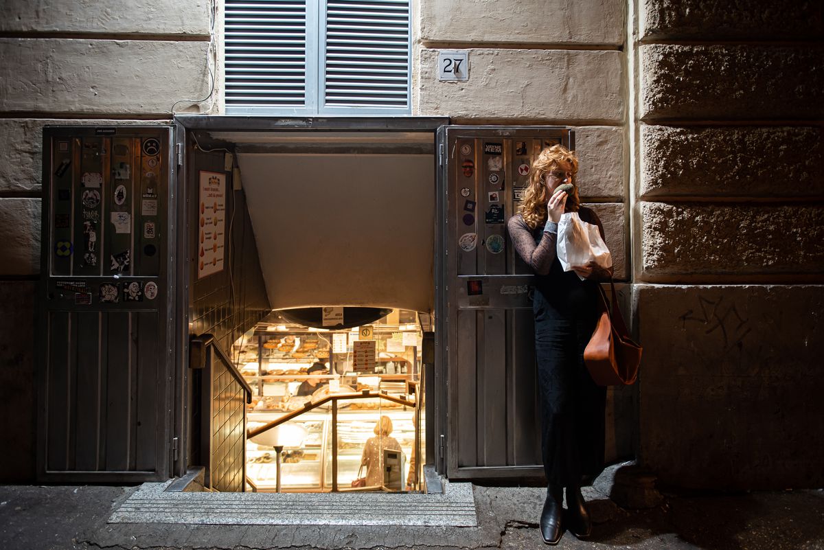 A woman stands alone eating a pastry, while leaning against the wall of a bakery. Light from the bakery is visible down a stairway beside her.