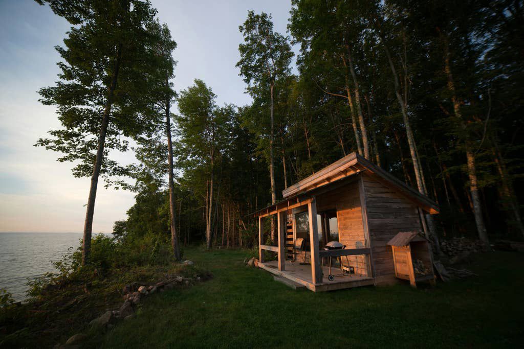 A wood cabin in a clearing on the waterfront of Lake Superior. The cabin has a front porch with a barbecue and chairs. There are trees in the back of the cabin.