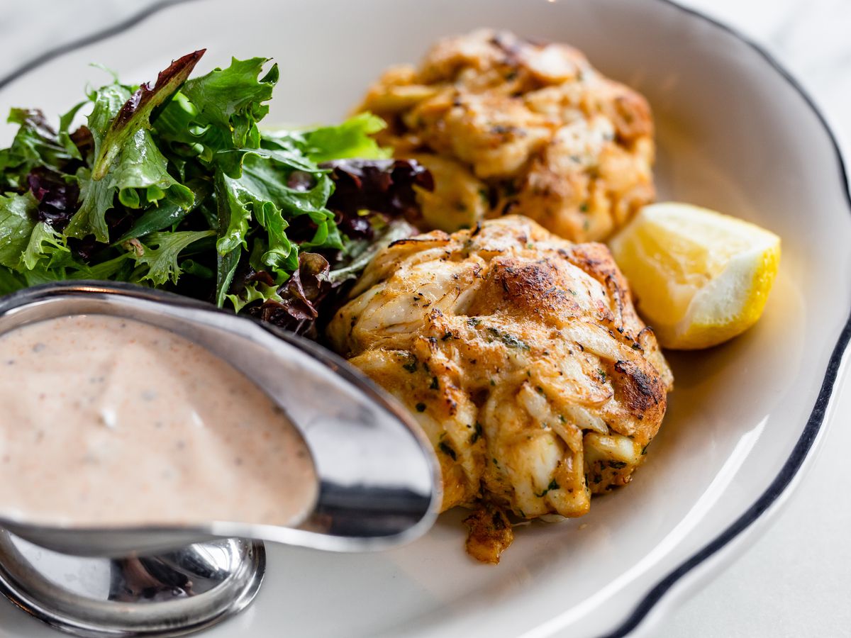 Two crab cakes with sauce on a white plate with blue trim, featuring a pink sauce on the side.