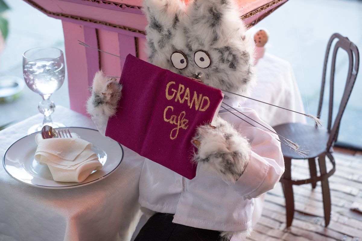 A gray and white cat puppet sits at a tiny table, reading a Grand Cafe menu