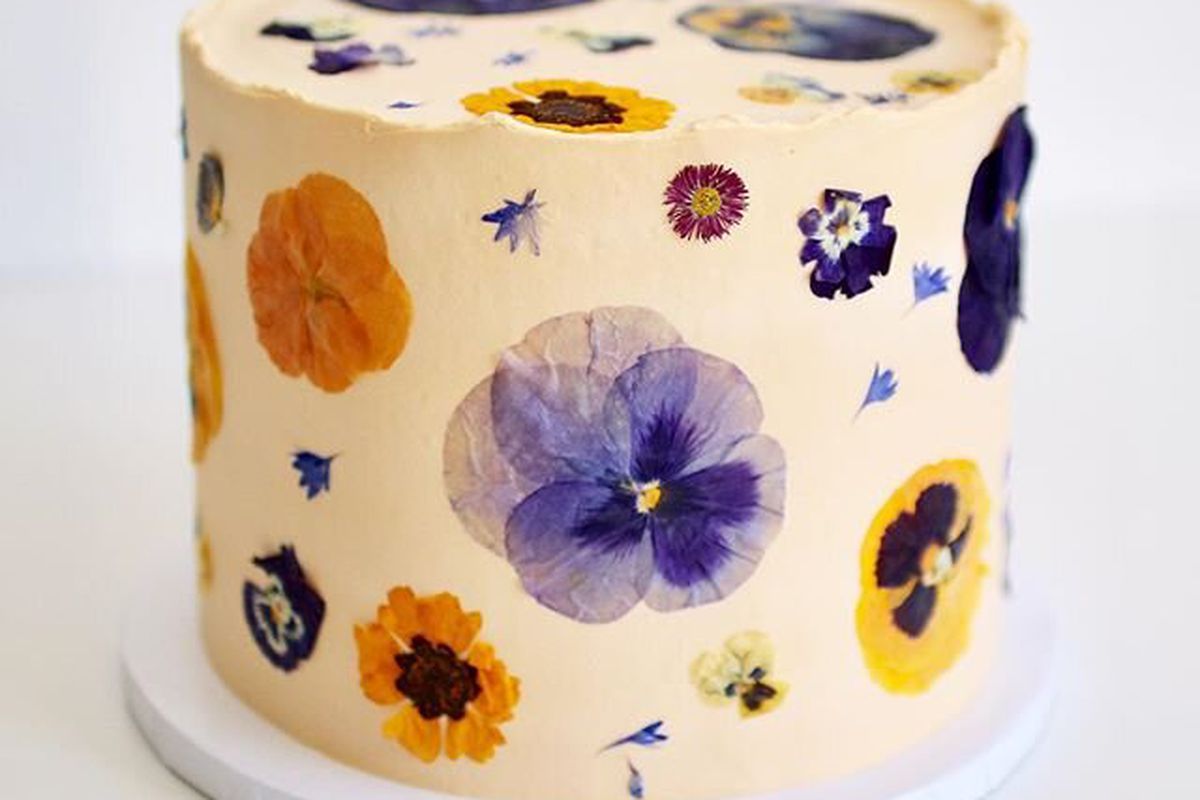 Orange and purple flowers of different sizes are pressed onto the surface of a cream-colored cake.
