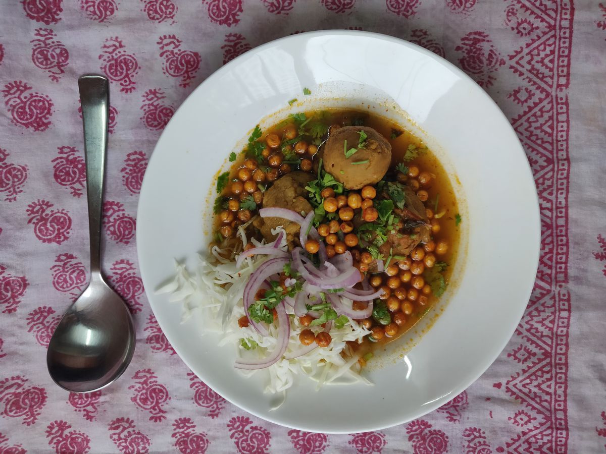 Seen from above, a bowl of curry with chickpeas, loose dumplings, onions, herbs and shaved cabbage, sitting on a decorative patterned tablecloth.