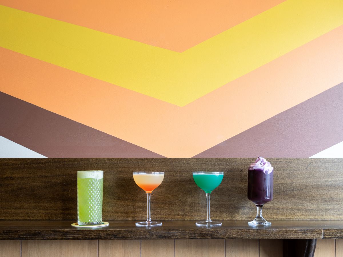 Four cocktails on a wooden bar rail in front of a retro mural on a wall.