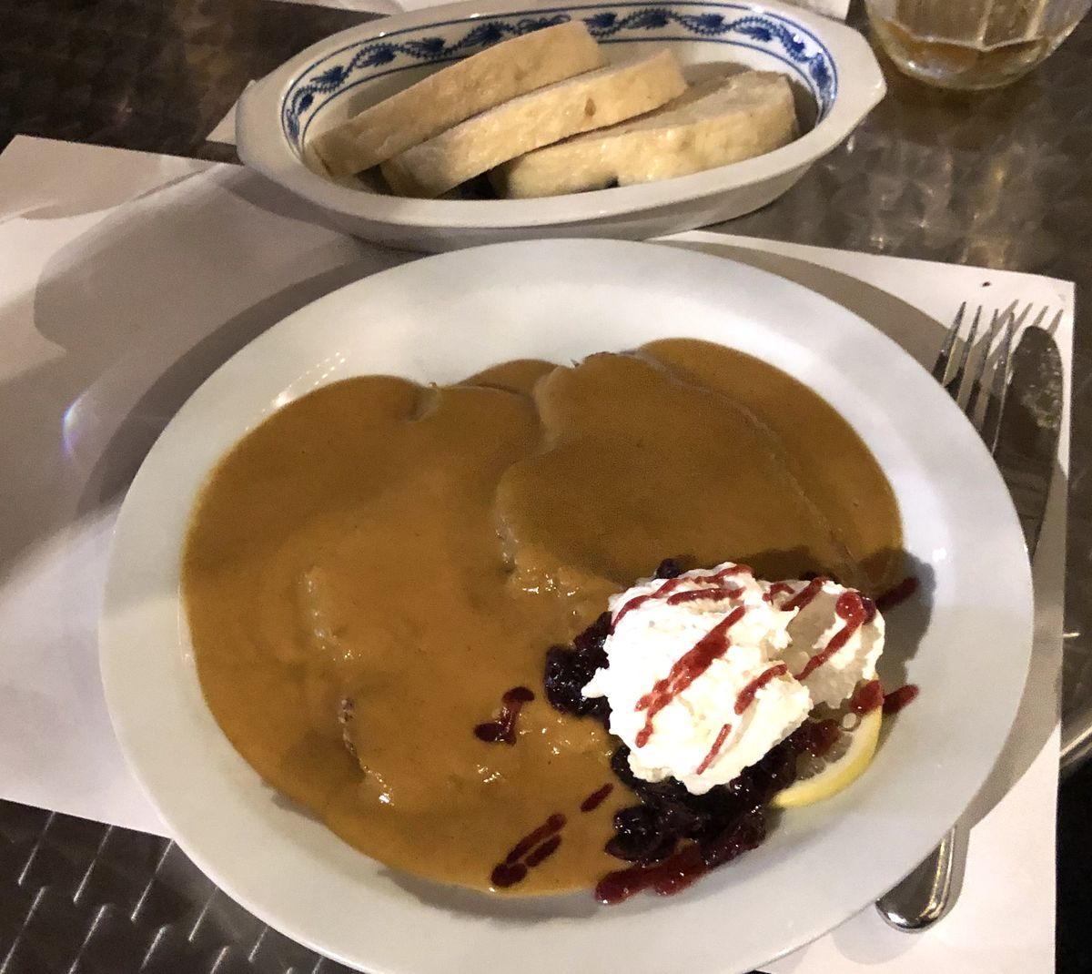 A plate of the Czech dish svickova, which has a brownish sauce and comes with a side of whipped cream and reddish berry-like jam and a slice of lemon
