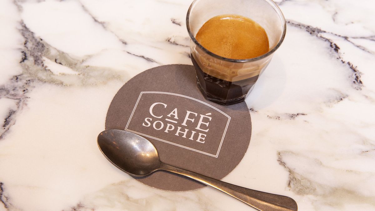 A round coaster with the words “cafe sophie” with a metal teaspoon resting and a shot of espresso in a glass.